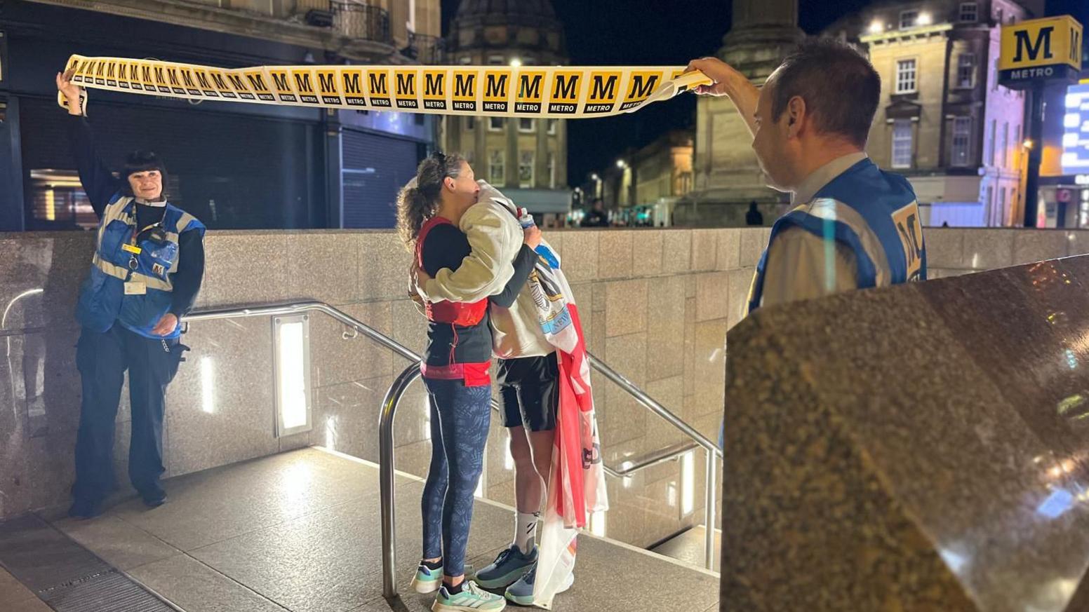 A teenager in running hear being hugged by a woman in a red jacket at the top of the steps of his final Metro station. 