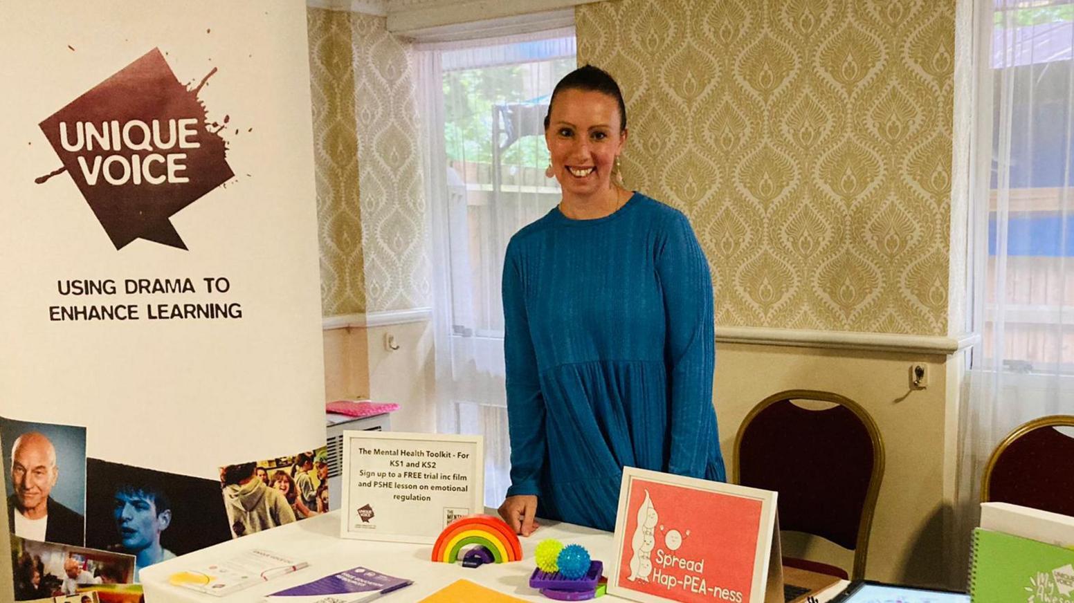 Krystal Keeley, wearing a blue long sleeved top, standing behind a table with interactive toys - a rainbow and stress balls - on. There is a Unique Voice banner to the left of the picture with a slogan, 'using drama to enhance learning'. Behind Ms Keeley there is a wall with cashew style wallpaper and two long windows with a light net curtain over. 