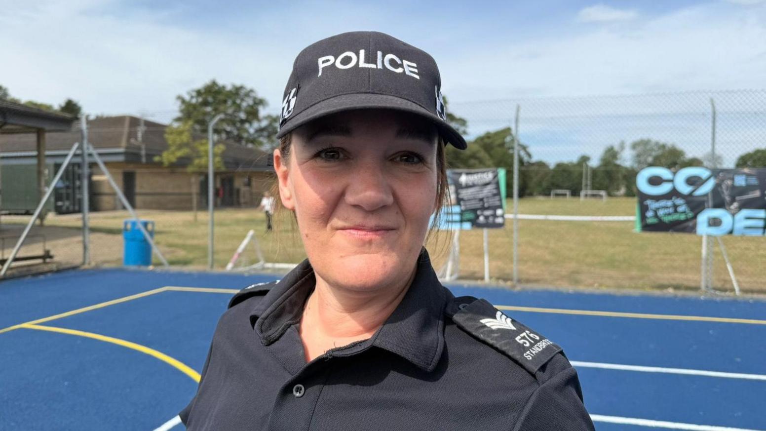 The photo shows a woman smiling at the camera. She's wearing a black baseball cap with the word POLICE written on it. She's also wearing police uniform. She's standing on a bright blue tennis court.