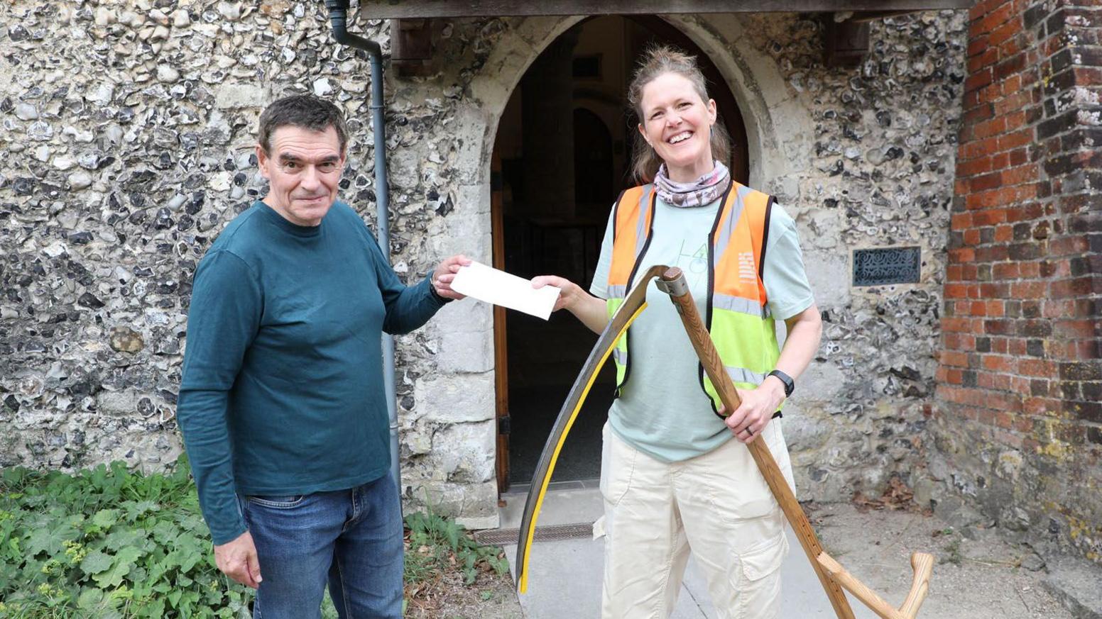 Church warden dressed in blue jeans and long sleeved top receiving check from a woman holding a large scythe and wearing cream cargo pants reflective sleeveless top. Both are standiung infront of church doorway.    