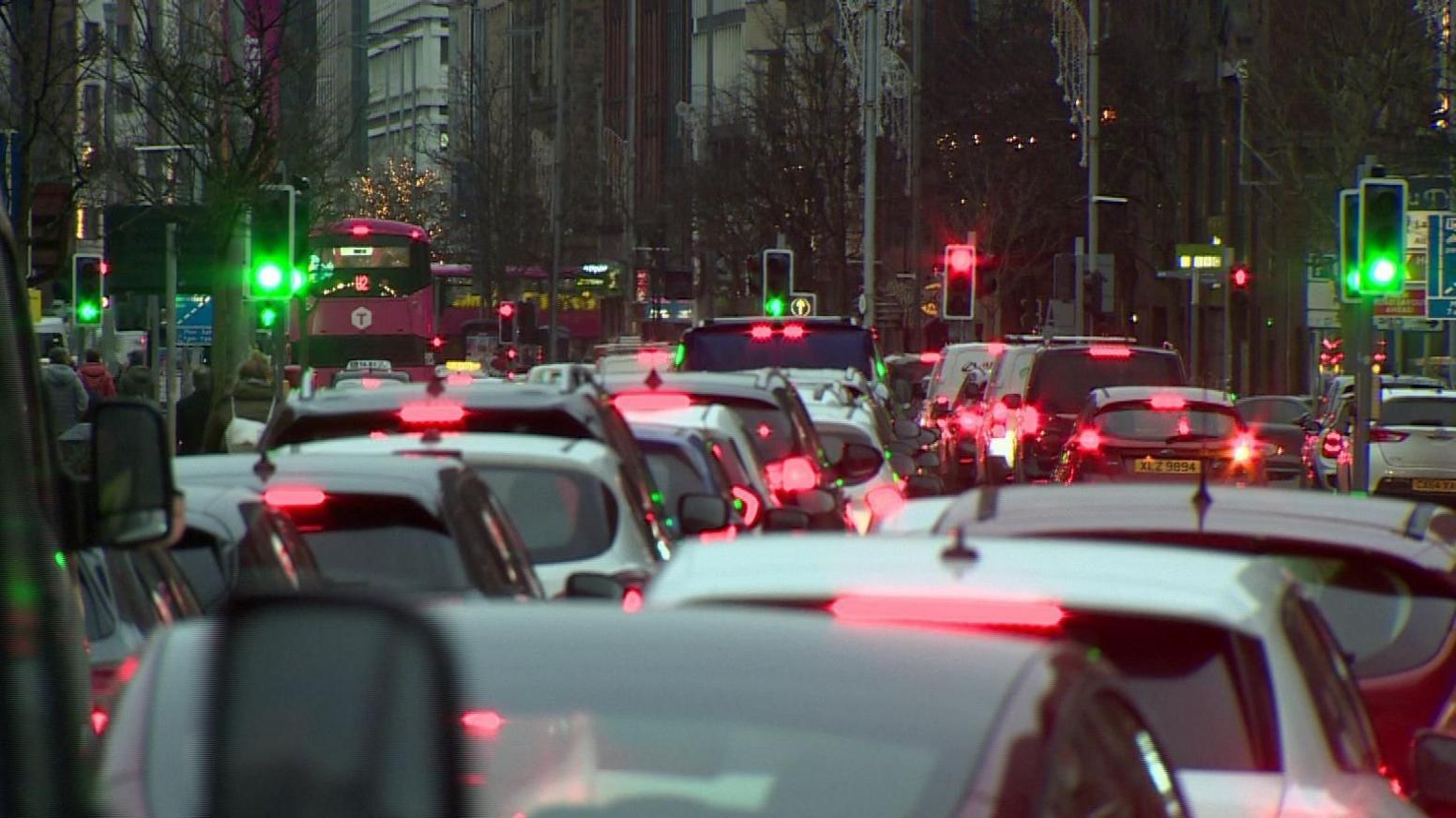 Backs of cars in traffic jam in Belfast city centre. Red break lights are visible and green and red traffic lights. There are trees lining the road and pink buses in the distance.