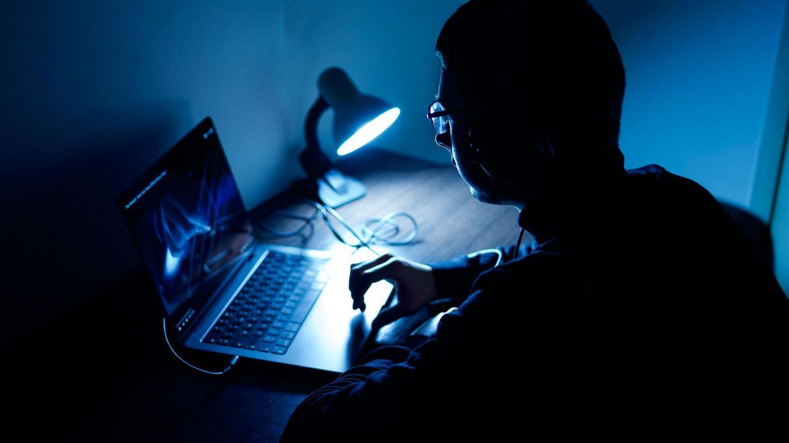 A man whose face is in shadow sits in front of a laptop