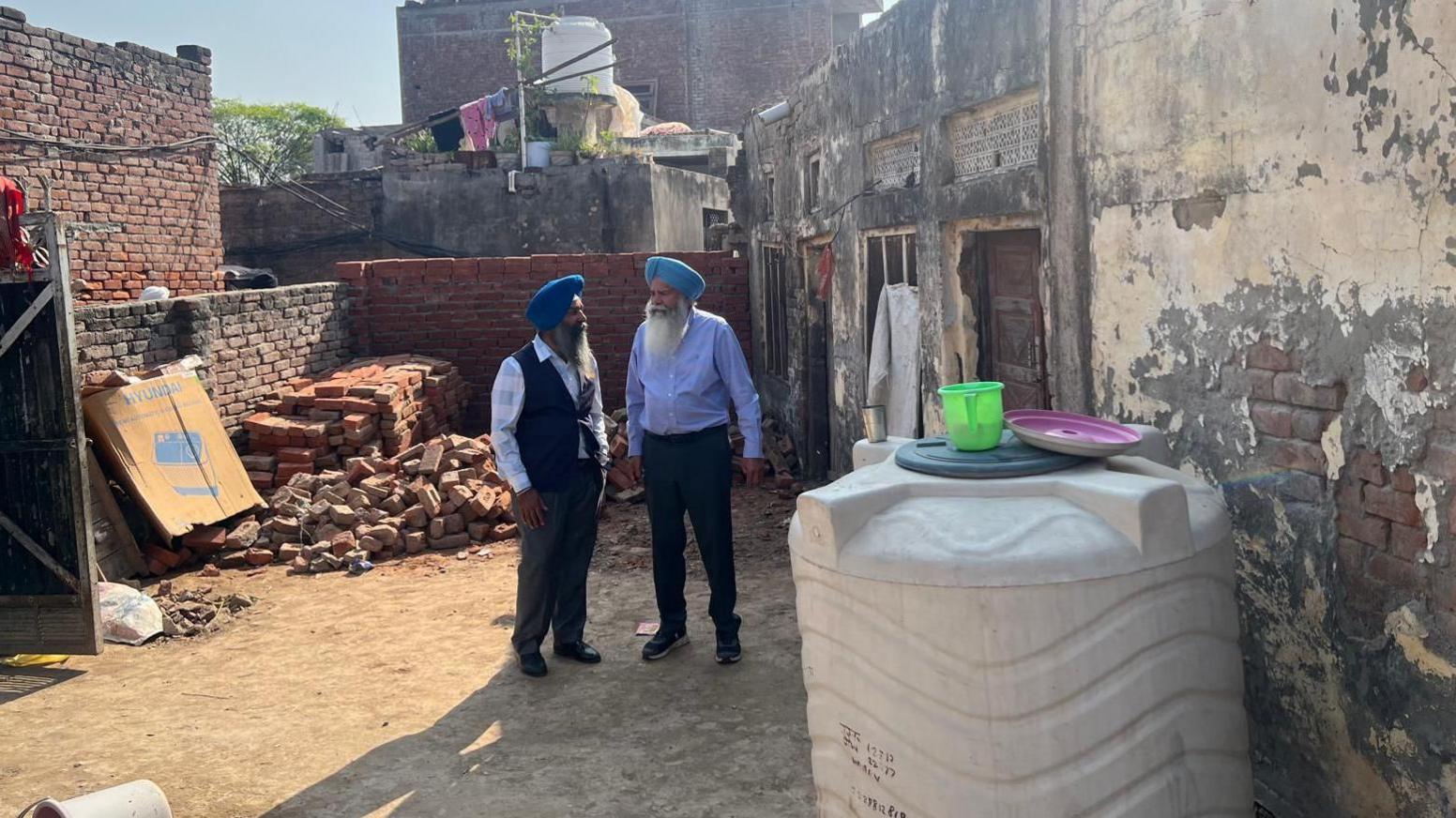 Two men in turbans and suits stand outside a dilapidated building with peeling paint and piles of bricks outside.