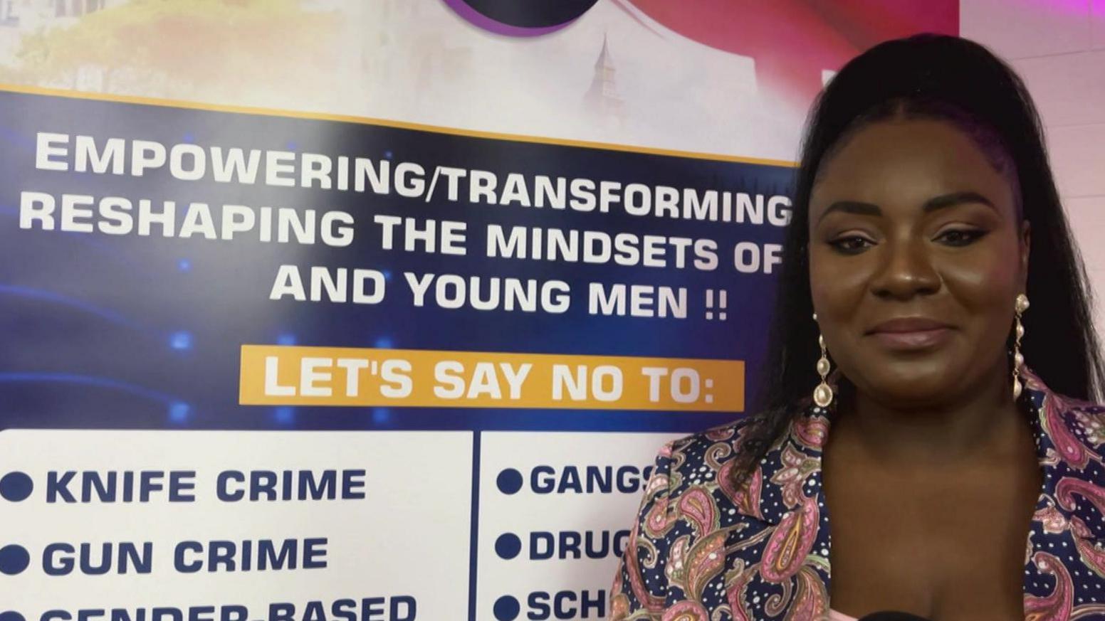 A woman wearing a pink and blue patterned suit stands in front of a banner promoting a youth project aimed at empowering young men and deterring them from crime and violence.