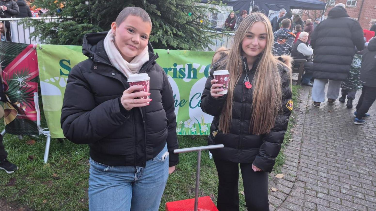 Izzy and Keely stand in front of a red box with a lever used to turn on Christmas lights before the event. The two friends are wearing black padded coat, holding disposable coffee cups and are standing in front of a Christmas tree which has a metal fence around it.