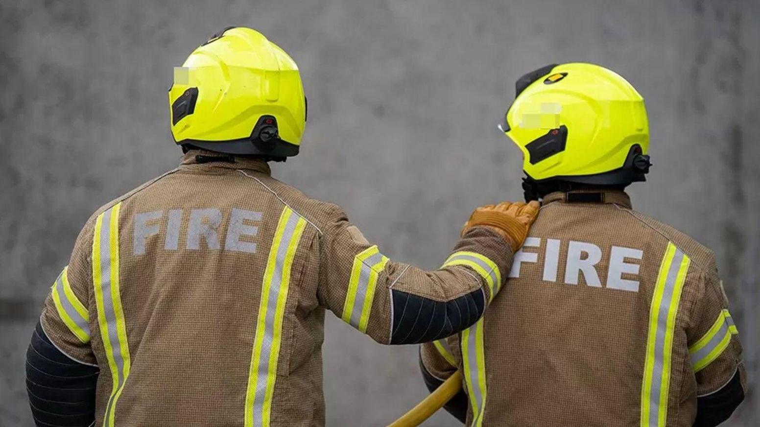 The backs of two firefighters wearing mainly brown uniform including helmets.