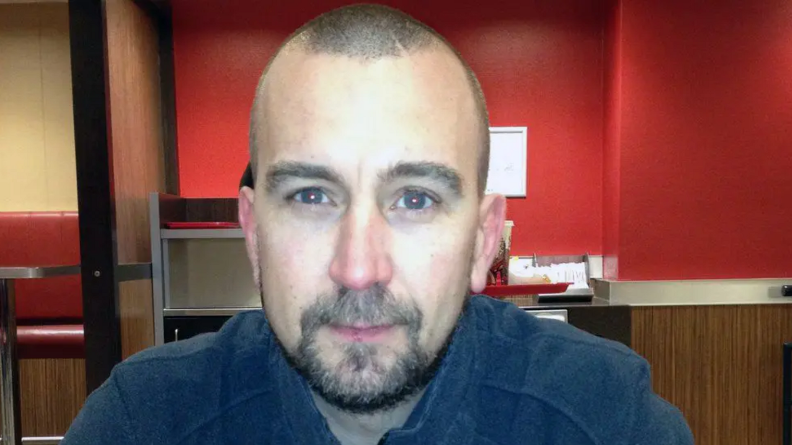 A headshot style picture of David Haines against a red background. He is looking at the camera. He has a shaved head and short beard.