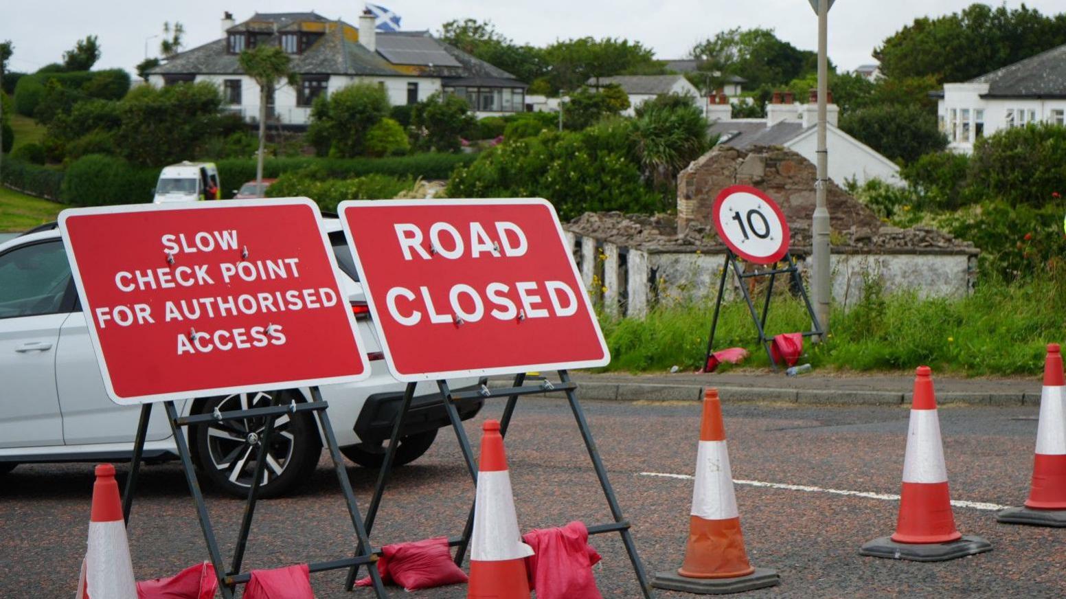 Traffic cones and three red and white signs block a road in a residential area. A number of white bungalows can be seen in the background. The rectangular signs read "SLOW CHECK POINT FOR AUTHORISED ACCESS" and "ROAD CLOSE". A circular "10" speed sign is also standing nearby. The signs are standing at an angle, as if they have been partially tipped over.
