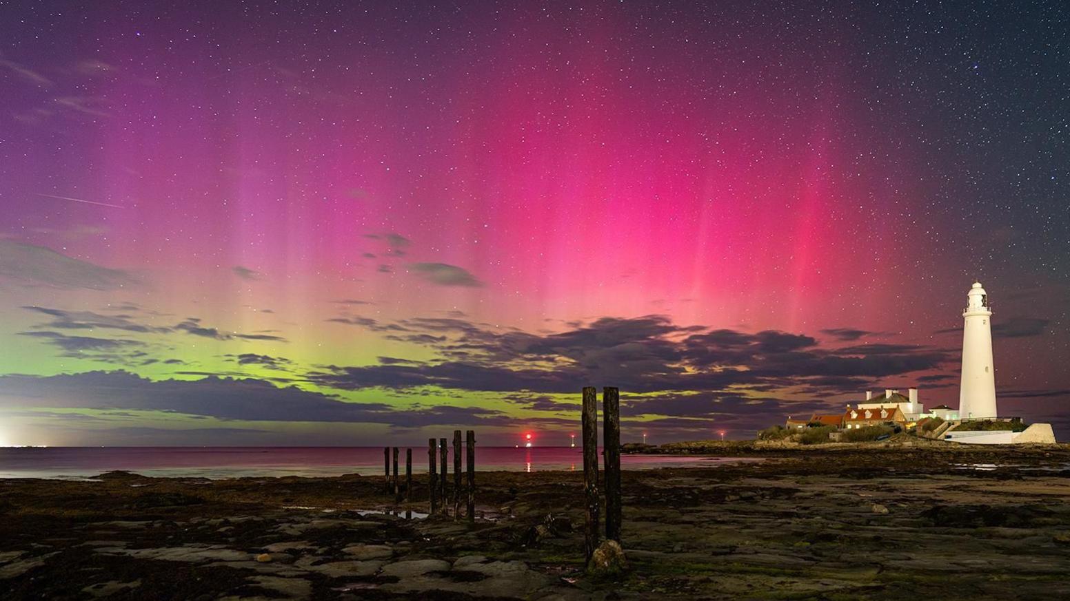 A view across a rocky beach as the tide is out, revealing old tall wooden beams sticking up. In the distance is a white light house which is clearly visible. The sea is far out. The night sky is lit up in green and pink, with lots of stars, as the northern lights are clearly visible.