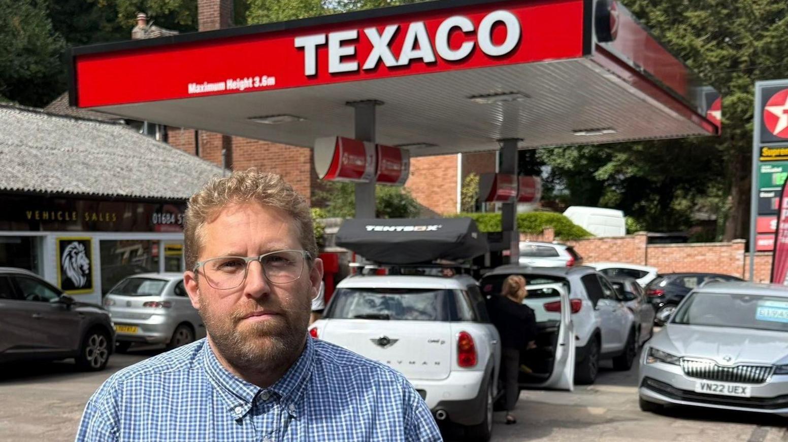 A man with a blue and white checked shirt wearing glasses. He has blonde hair and has a beard. He is standing infront of a car dealership