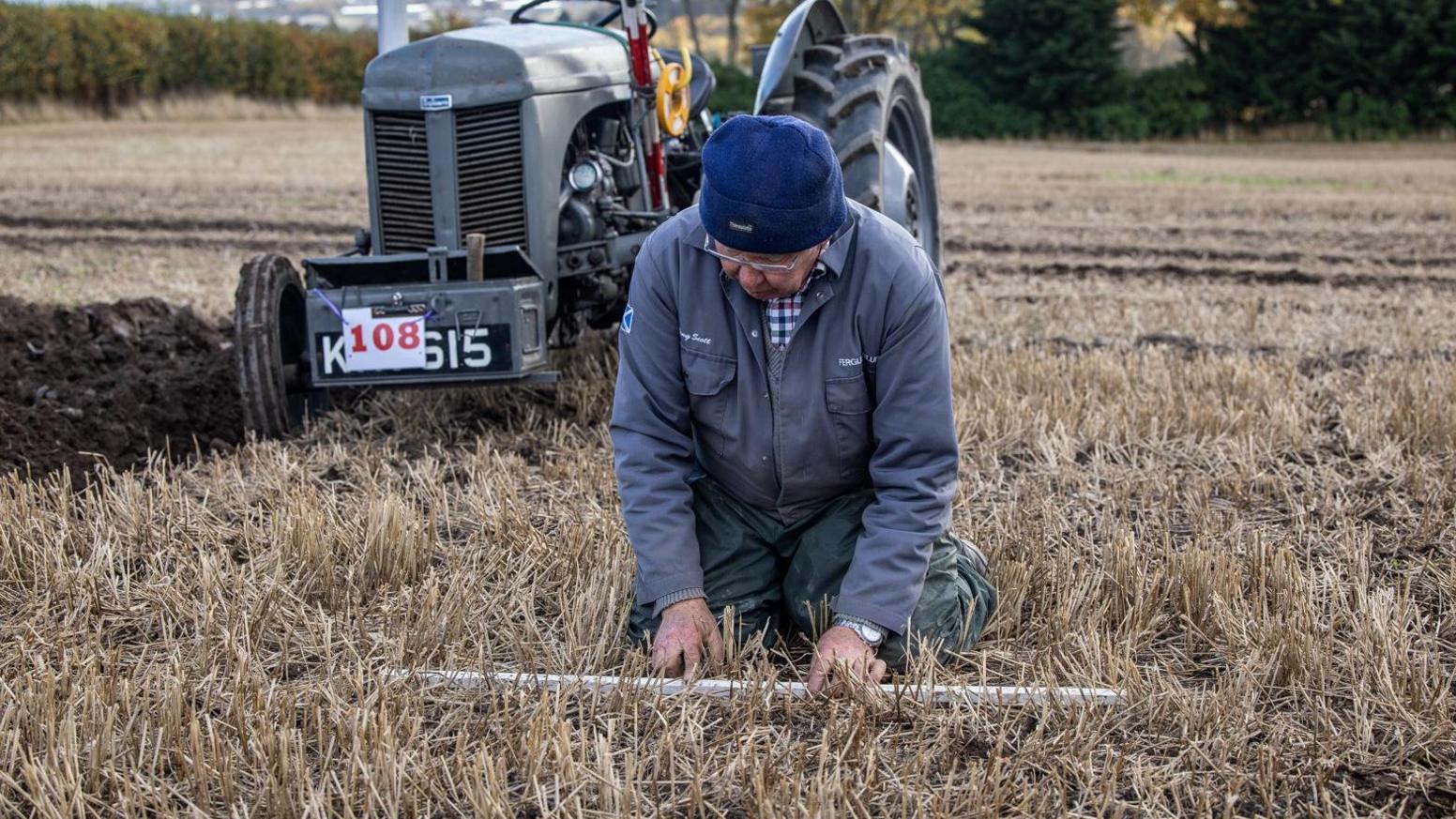 A competitor plans his ploughing. He is wearing a grey boiler suit and blue woolly hat. His grey tractor is in the background.