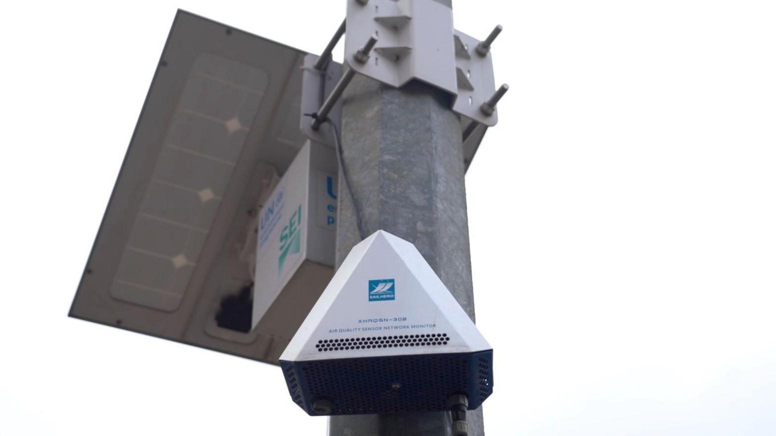 A triangular air quality monitoring sensor is affixed to a post, with wires leading to a box topped with solar panels