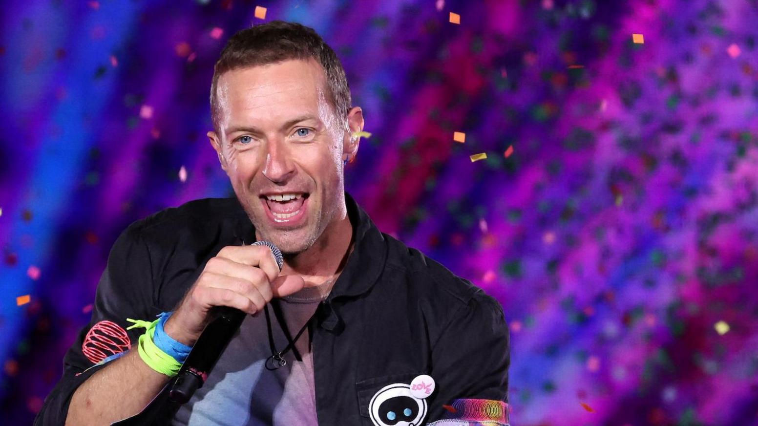 Chris Martin of Coldplay singing on stage with a colourful background