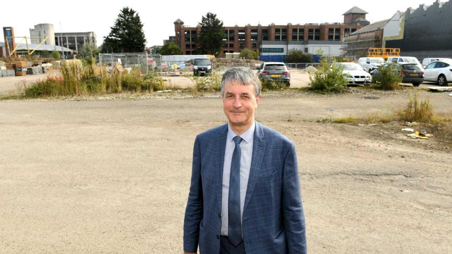 Philip wears a blue checkered suit and blue tie and is standing on the brownfield site which backs on to a car park.  There are buildings in the background and metal barriers with bits of rubbish blown into them.