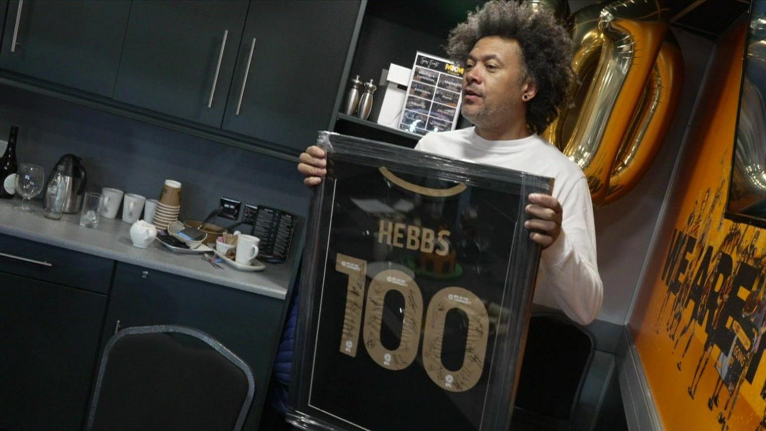 Former footballer Jason Price holds a framed souvenir shirt in an executive box at a football stadium. Mr Price has dark brown hair and wears a white T-shirt. The shirt is black with gold details, including the name "Hebbs" and the number "100". Signatures can be seen written in black pen on the golden numbers.