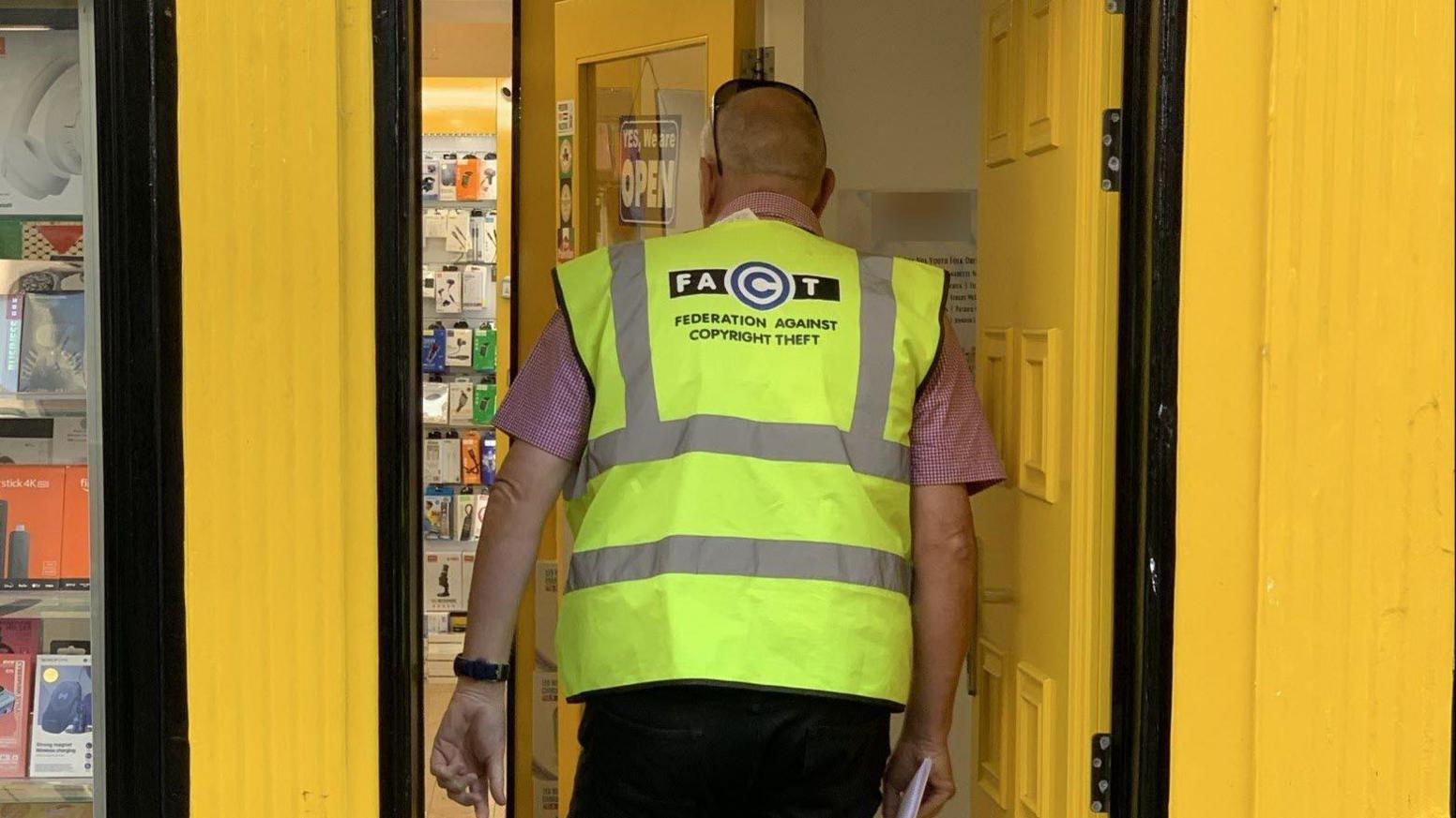A man enters a yellow shop front wearing a Fact hi-vis jacket. He is also wearing a pair of sunglasses on his head, and a short-sleeve check shirt. He has his back turned to the camera.