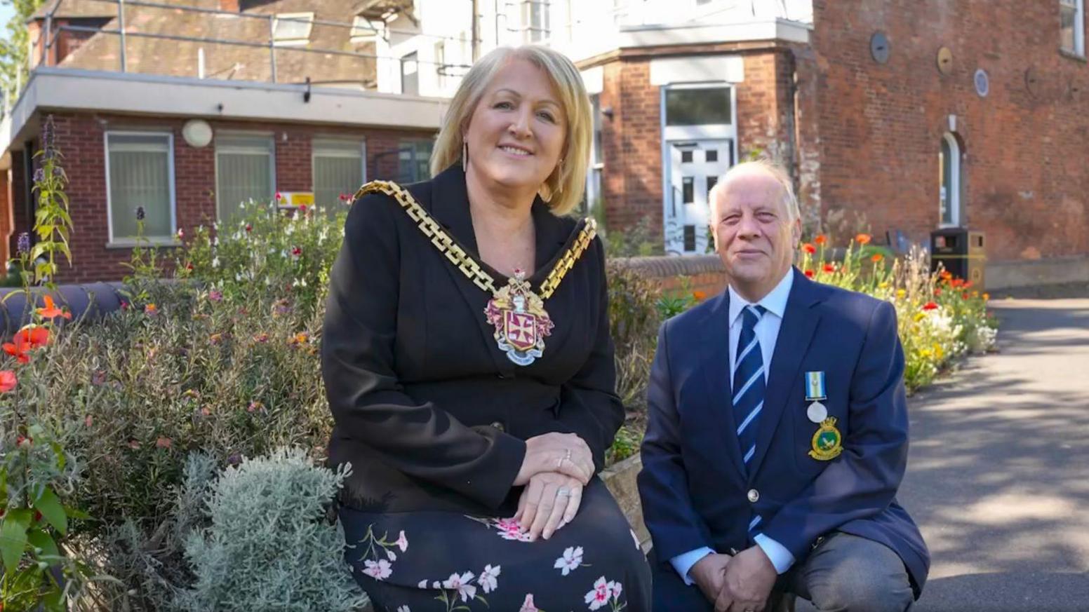 A woman wearing a mayoral chain sits on a low garden wall with her hands clasped, next to a man crouched next to her wearing a dark blue suit and a military medal. Behind them is a row of plants in front of a building.