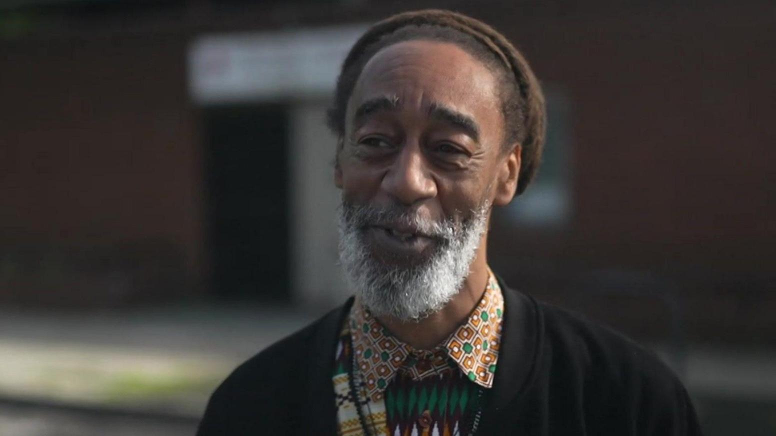 Man with grey beard wearing orange and green patterned shirt