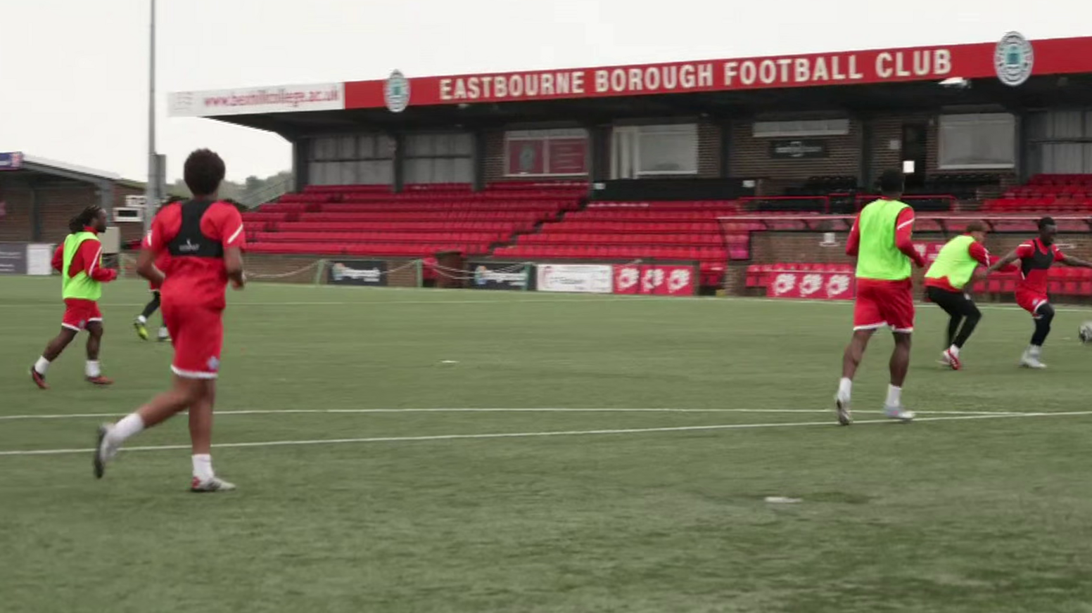 Players on a green football pitch. All are wearing red kit and some have neon green bibs on. A short stand with red seats in the background has Eastbourne Borough Football Club written across the top.
