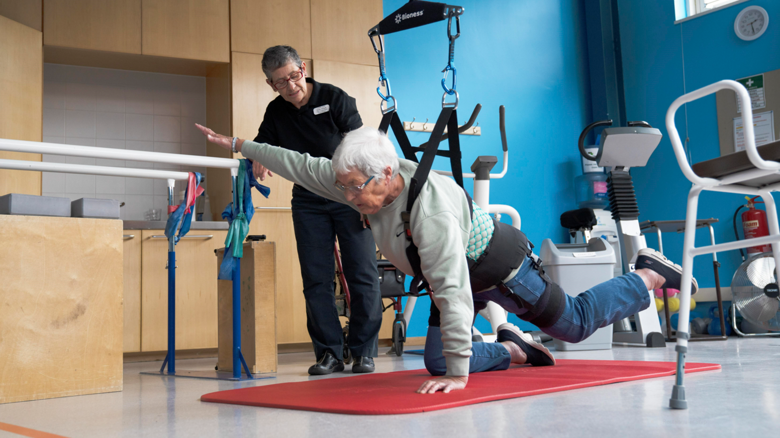 A woman supported by a ceiling harness poses on a yoga mat on all fours, with her right arm and left leg raised and outstretched, watched by a woman in dark clothing whose name badge on her chest in not readable.