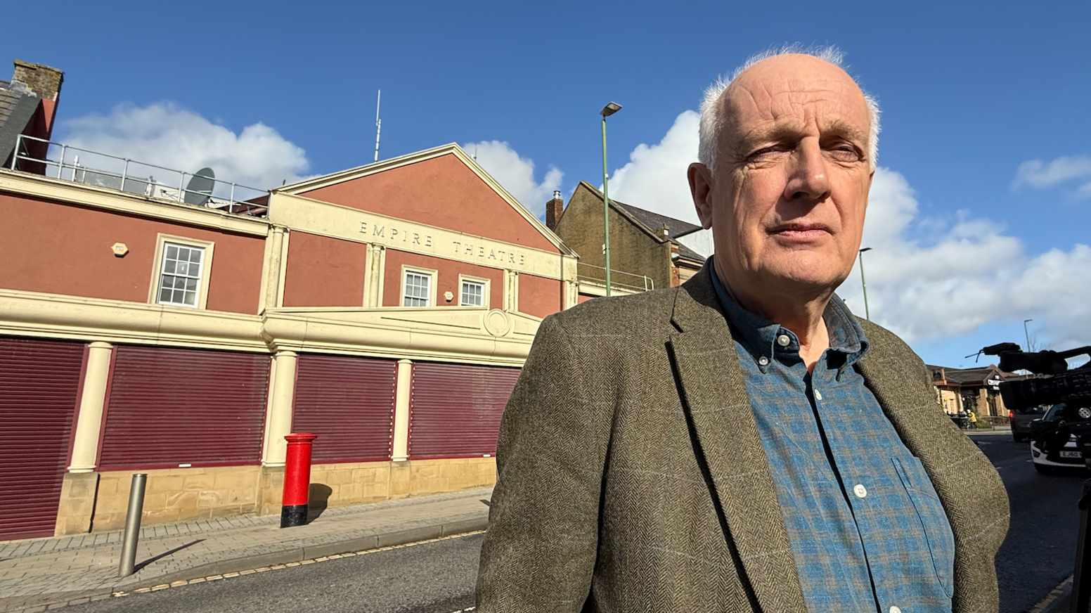 Kevin Earley has white hair and is wearing a tweed style jacket over a blue and grey check shirt. He stands in front of the Empire Theatre building, which has red brickwork and yellow/beige render. Maroon shutters are closed in front of the door and ground floor windows.