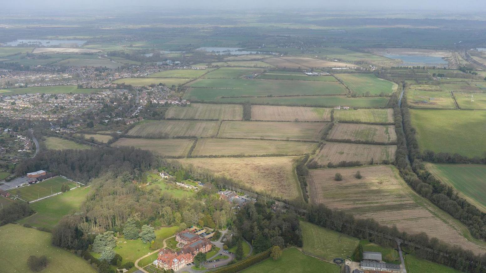 An aerial view of green fields with a railway line on the right and houses on the left. At the bottom there is a large property surrounded by trees.