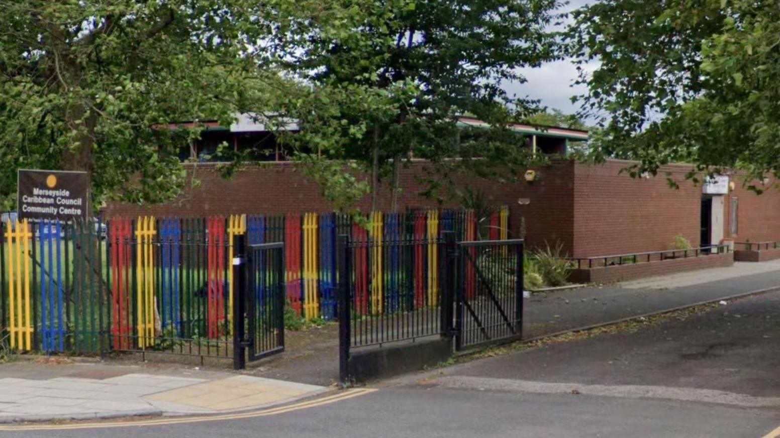 A single storey brick building with red, yellow, blue, black and green painted railings. A brown sign with white writing reads: Merseyside Caribbean Community Centre