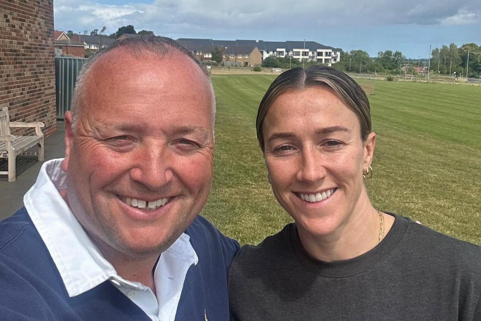 Head-and-shoulders selfie of Ryan Jobson (left) with Lucy Bronze. They are both looking into the camera and smiling. Mr Jobson has short grey hair and is wearing a navy rugby-shirt while Bronze has her hair tied back is is wearing a dark jumper. A sports pitch can be seen in the background.