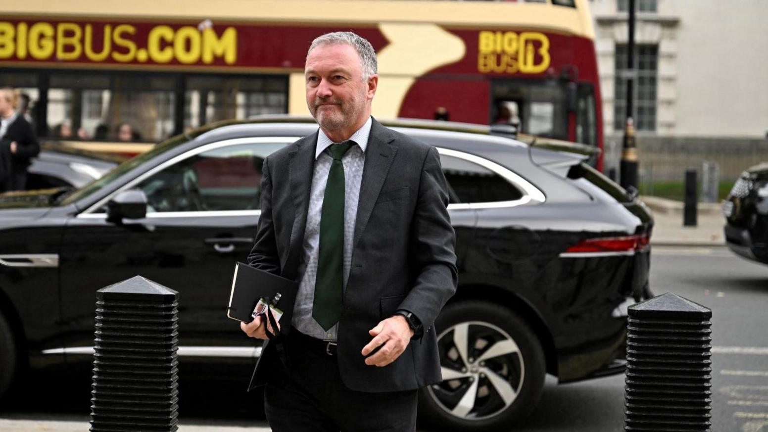 A man in a dark suit and tie carries a book and a security pass as though entering a building