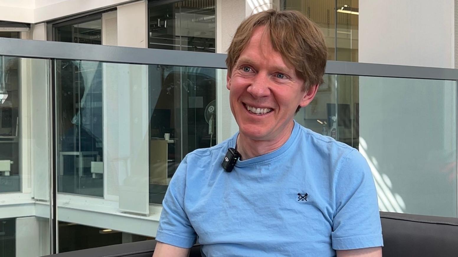 Peter Bell smiling widely and happily while looking to the side - he is wearing a light blue shirt and has blonde mid-length hair.