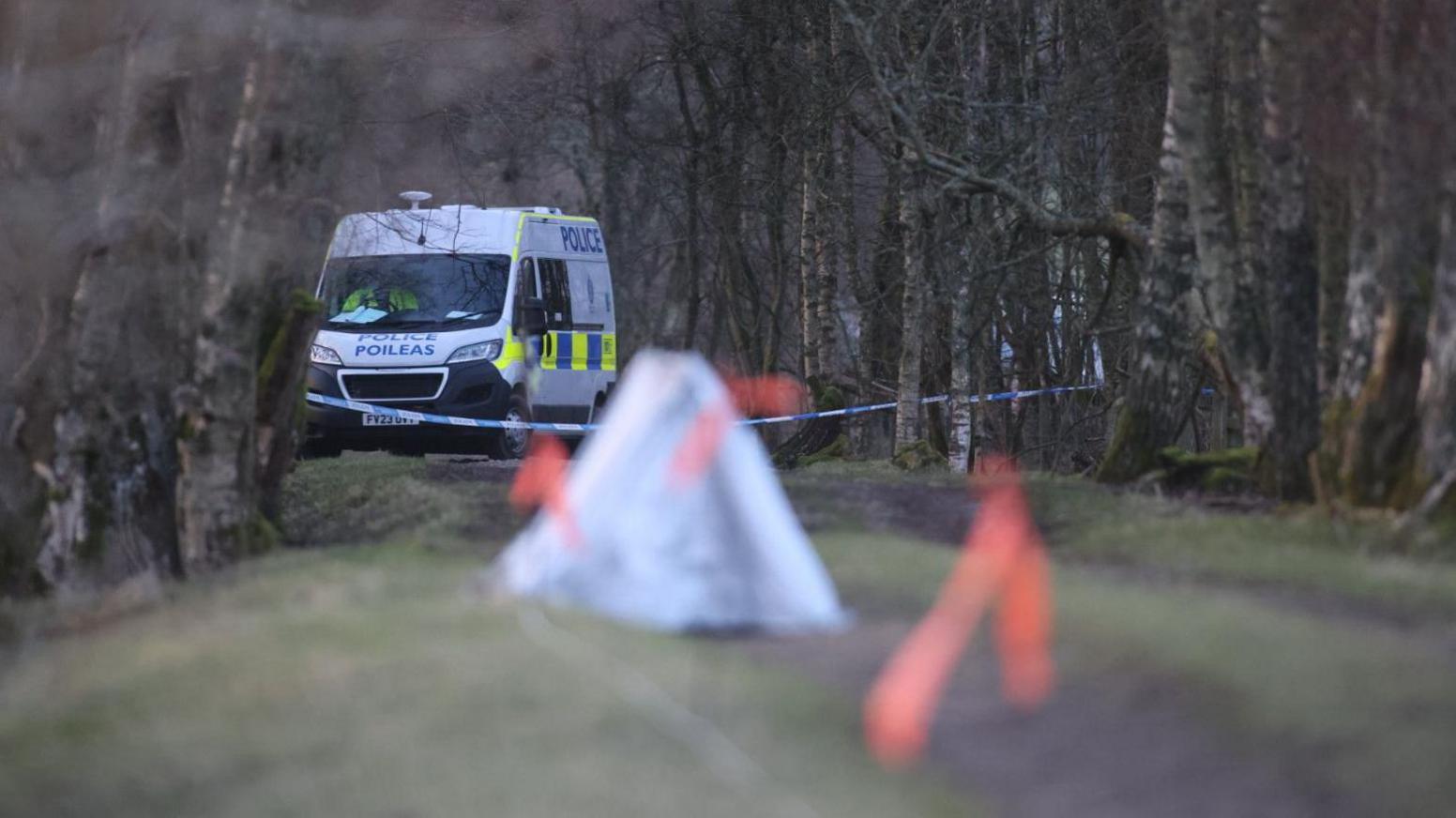A police van sits on a rural path with police tape, a white forensics tent, and orange markers in the ground