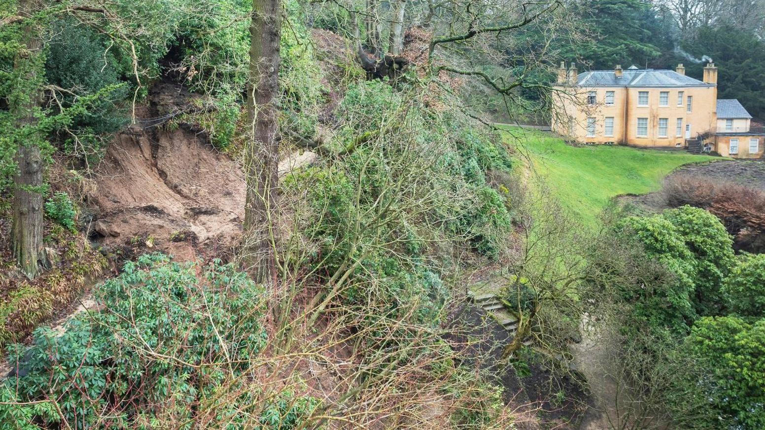 Fallen shrubs and the impact of landslides can be seen in the estate's garden. A large building can be seen in the background.