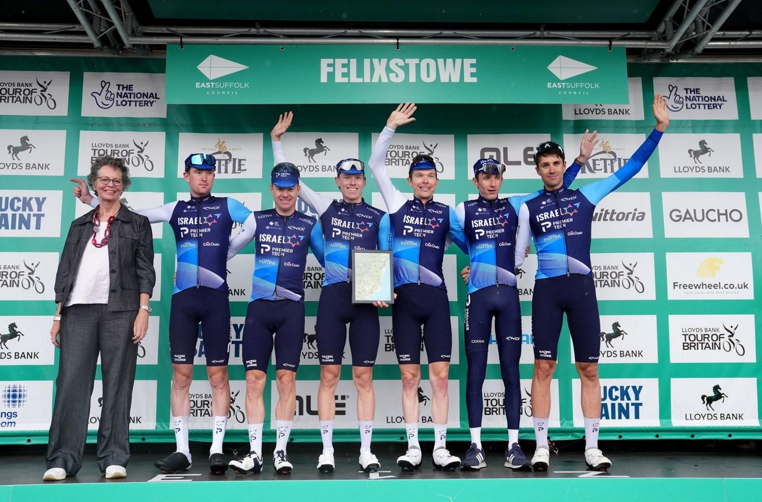 Six men in the Israel Premier-Tech sponsored blue lycra cycling outfits stand on the podium and wave, with a lady on the left wearing glasses and grey suit