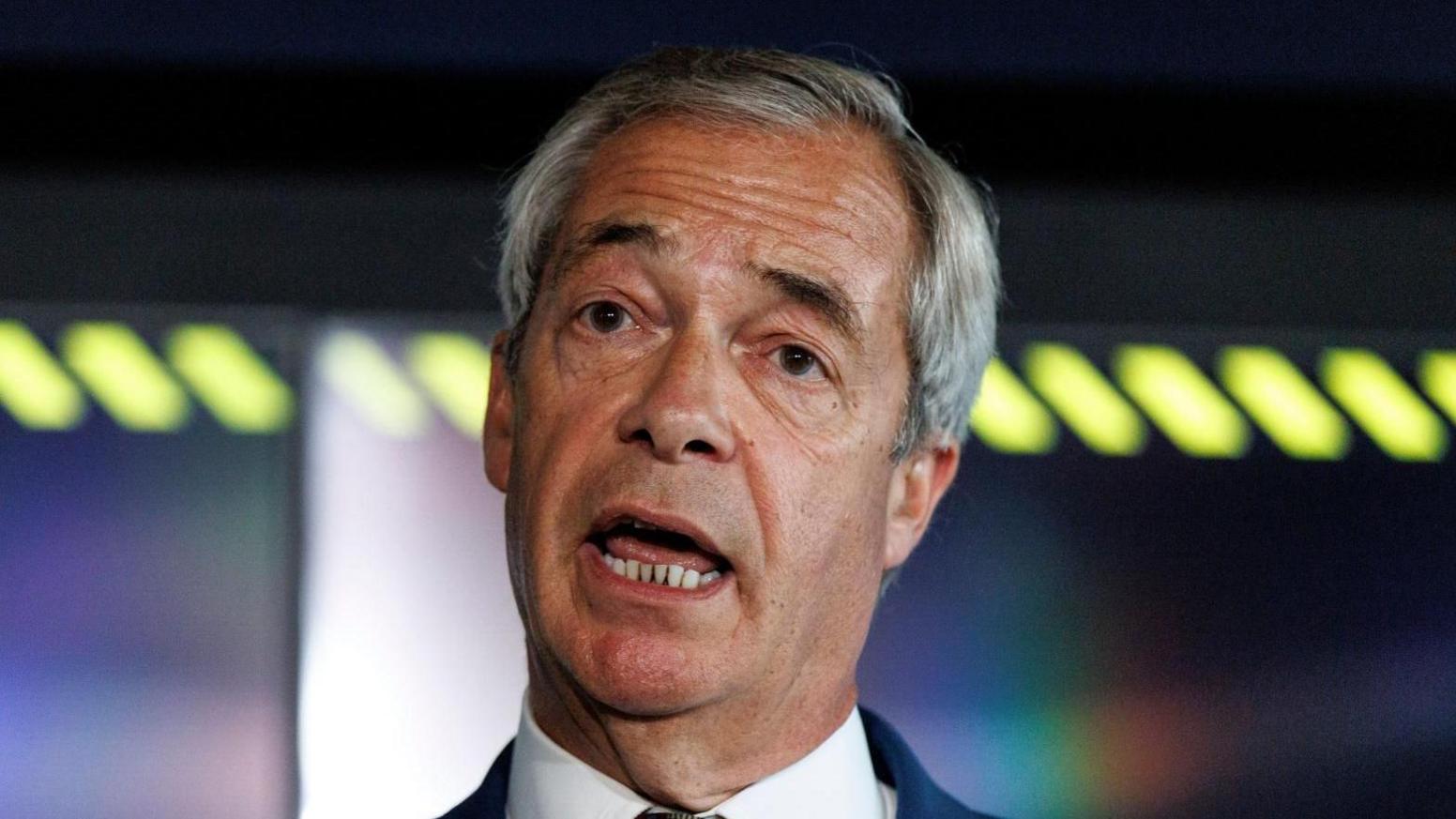 Nigel Farage, wearing a white shirt and navy blazer, tilts his head to one side as he speaks
