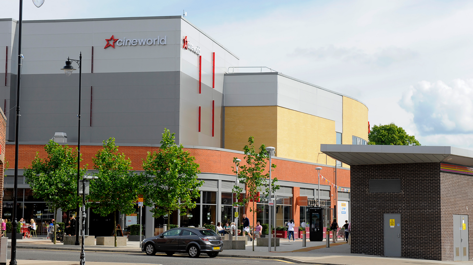 A Cineworld complex behind a row of trees and cars and pedestrians in the foreground 