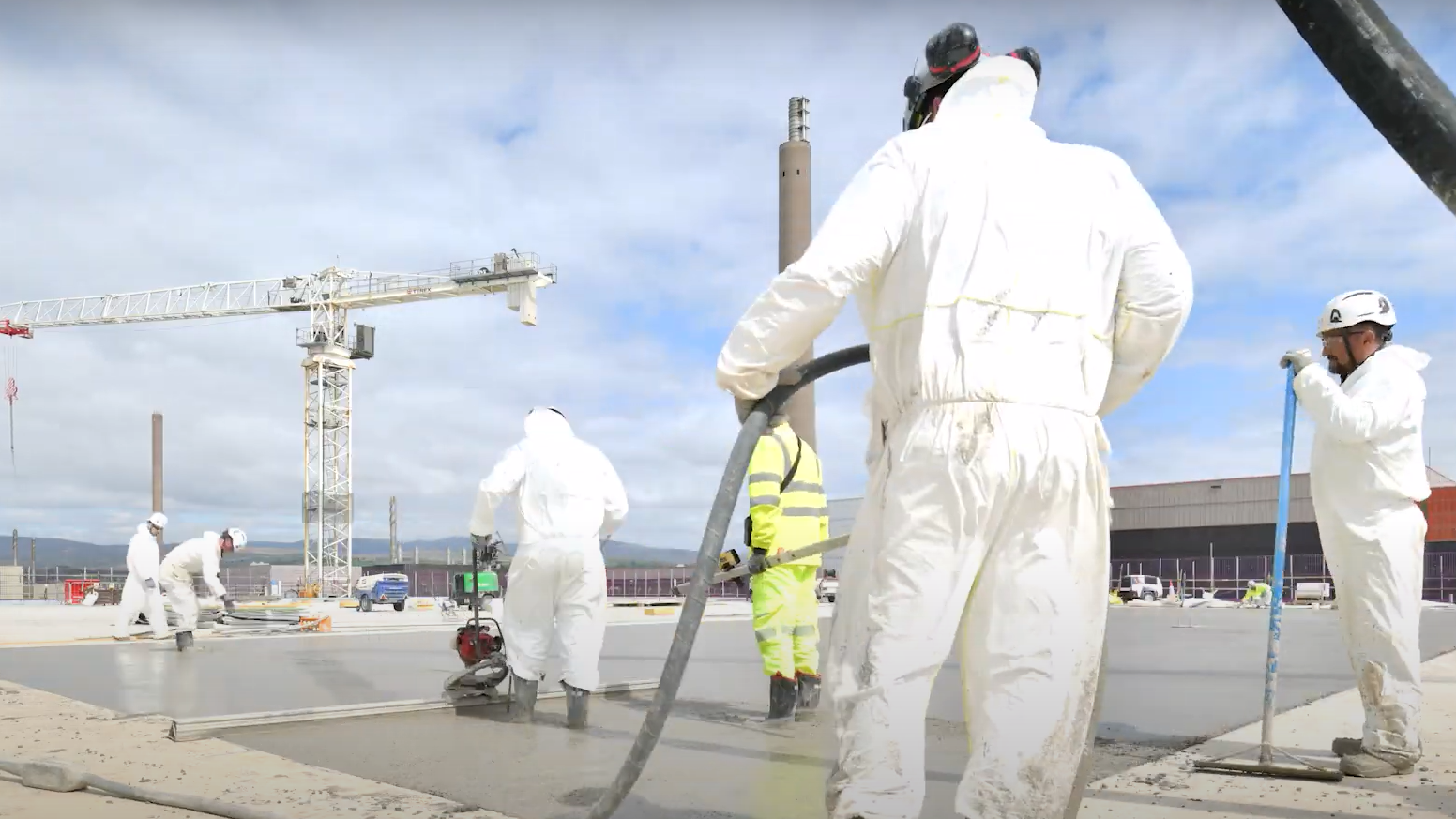 Workers wearing white suits and florescent overalls lay concrete at the treatment plant in Sellafield. There is a crane in the distance and other industrial buildings.