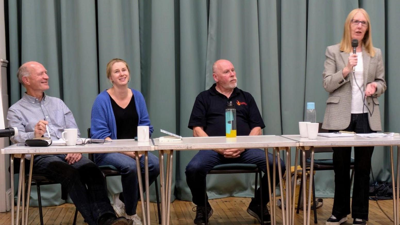 Four people sit at a table at a public meeting. Three are seated and one woman is on her feet giving a speech 