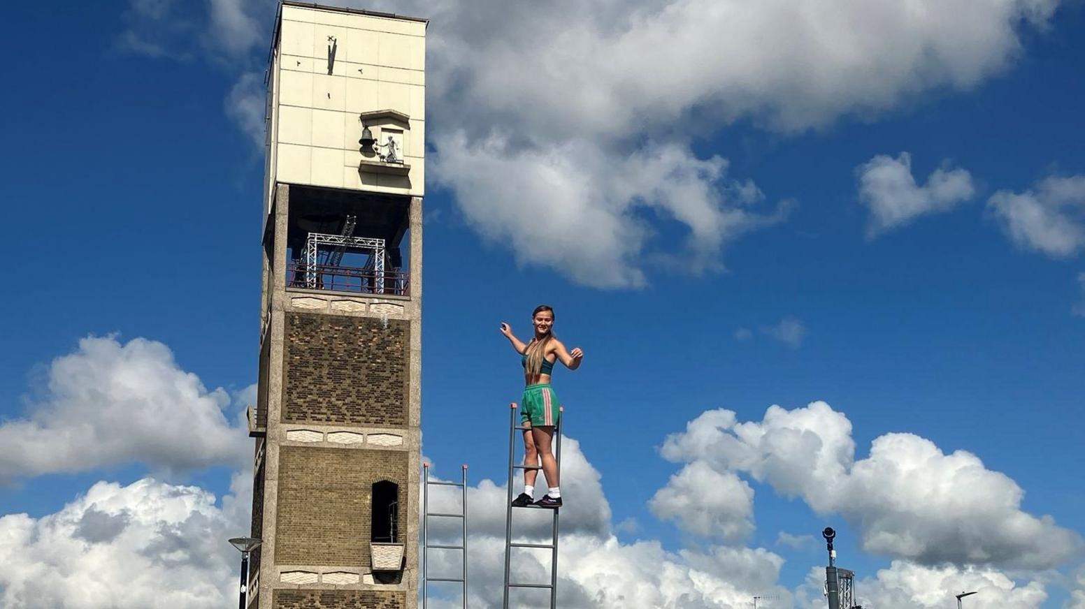 A circus performer balancing on a ladder next to a makeshift tower. She is wearing green shorts and a blue sports bra