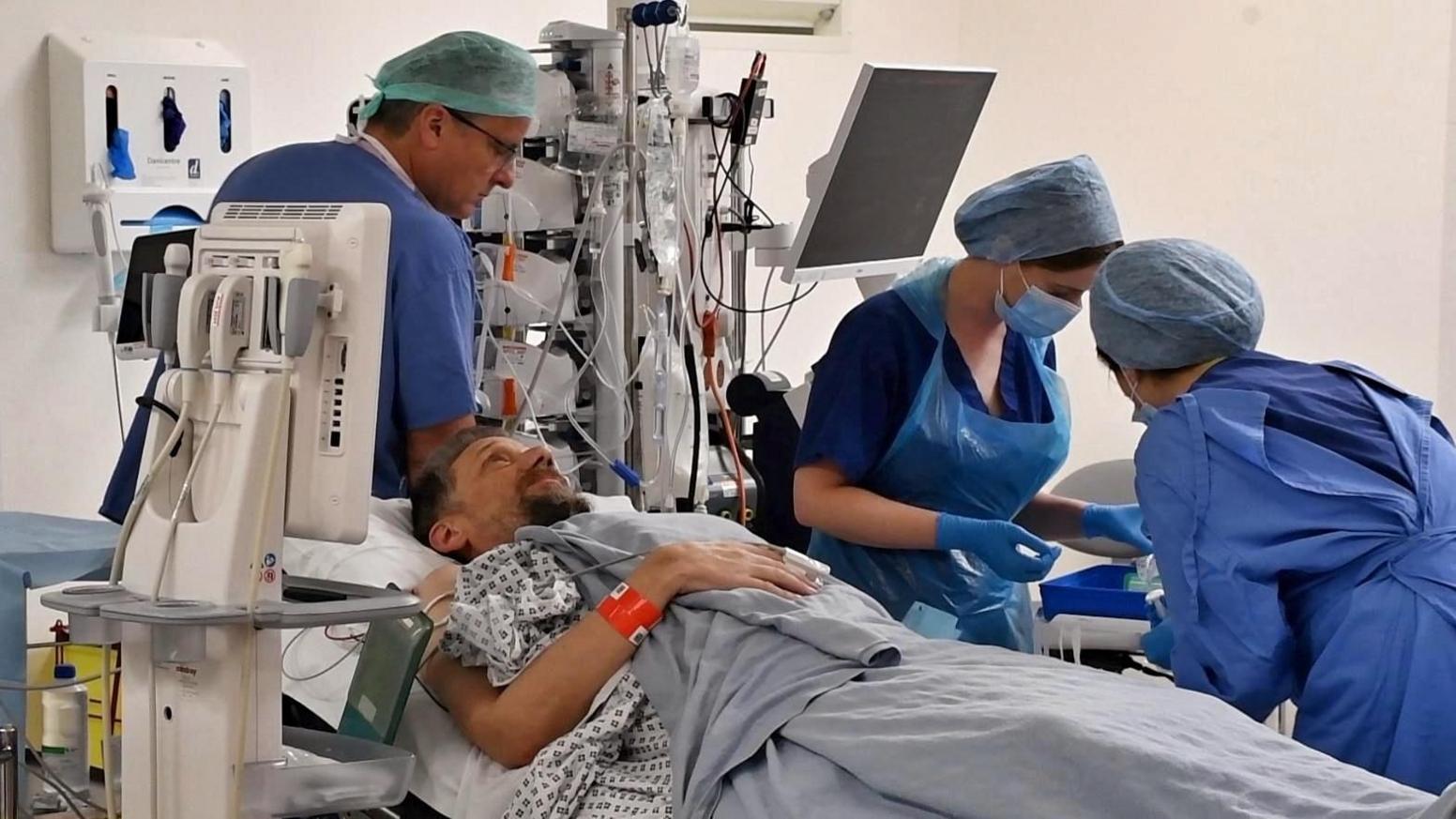 Daniel Evans-Smith lying on a bed in the operating theatre at Royal Papworth hospital. He has his eyes open and a wristband on and is looking up at the ceiling. A surgeon stands behind him wearing a hair net and scrubs. Two nurses are checking equipment to his left and are wearing scrubs and aprons