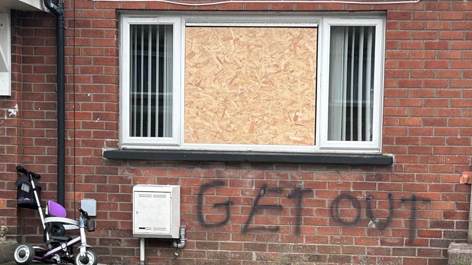 Close up picture of the spray painted "get out" beneath a boarded up window. There is a child's bike beside it.