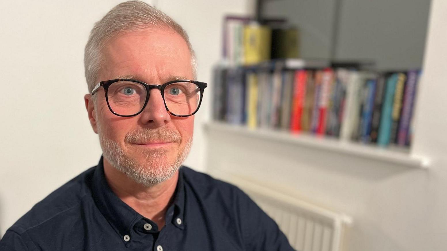 A man with light grey hair and a trimmed beard looks at the camera. He is wearing thick rimmed, round black glasses and and dark blue shirt with buttoned-down collar and an open neck. In the background is a shelf of books.