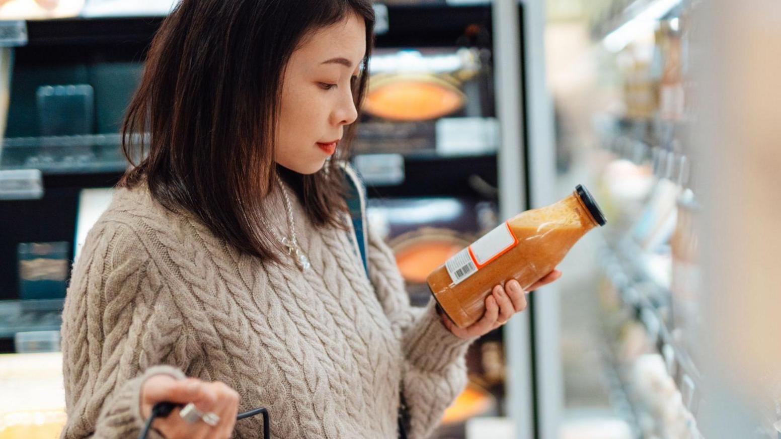 Young Asian woman in a jumper looking at a large juice bottle she has taken from a supermarket cabinet