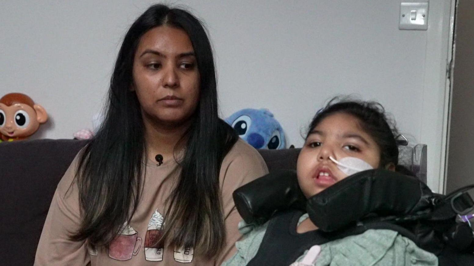 A picture of mum Namla (left) with dark brown hair and daughter Sara (right) sitting in a wheelchair with a feeding tube in her nose.