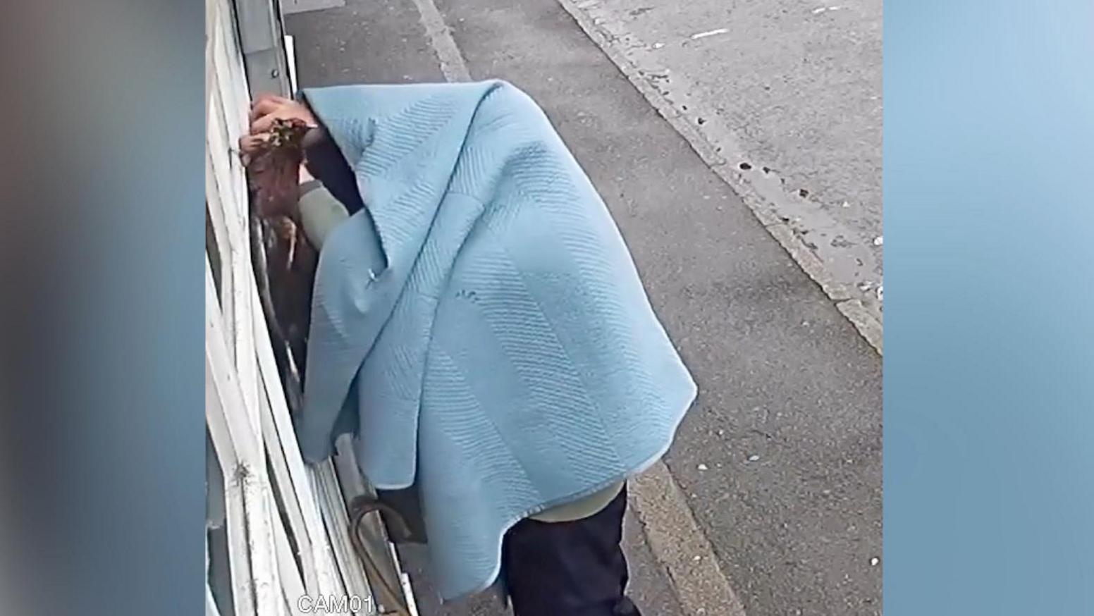 CCTV image of human figure, the upper half of whom is covered with a blue blanket, hanging two dead pheasants on the shop front