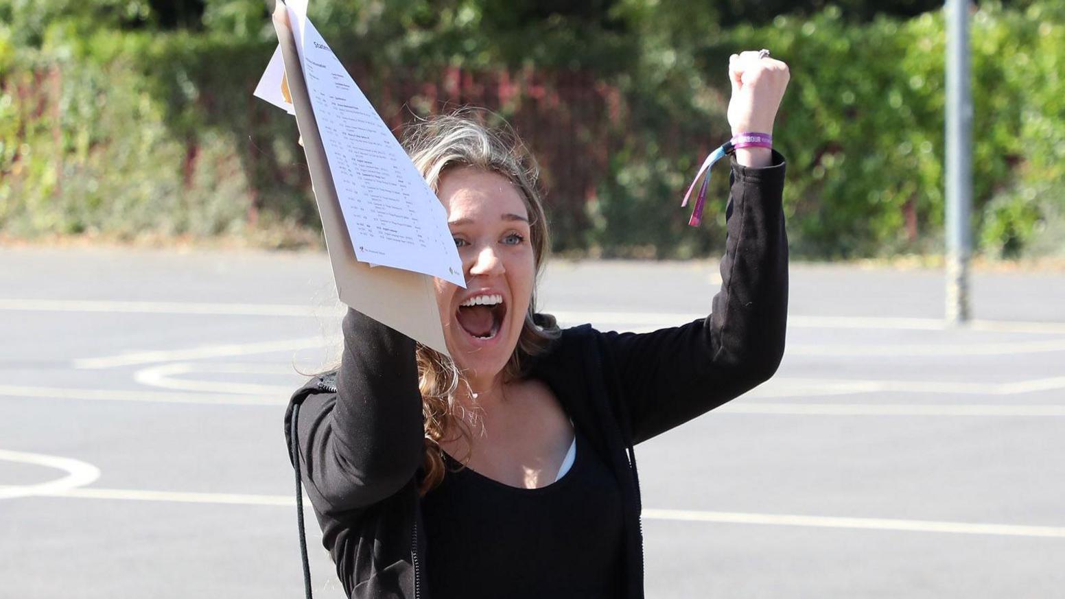 A young woman stands in an empty basketball court outside in her school grounds. She has her hands clasped in fists raised above her head and her mouth open like she's celebrating. She has dark blonde hair and wears a black zip hoodie with a black t shirt underneath. She is holding an open brown envelope and white piece of paper in her right hand.