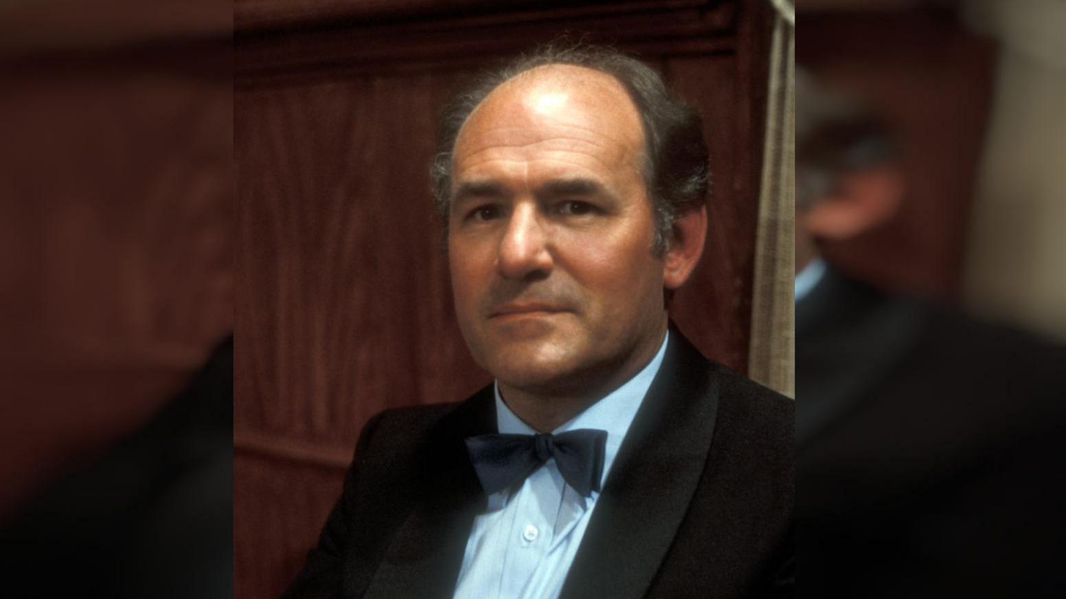 A bald man in a black suit with a bow tie. He is standing behind dark wood.