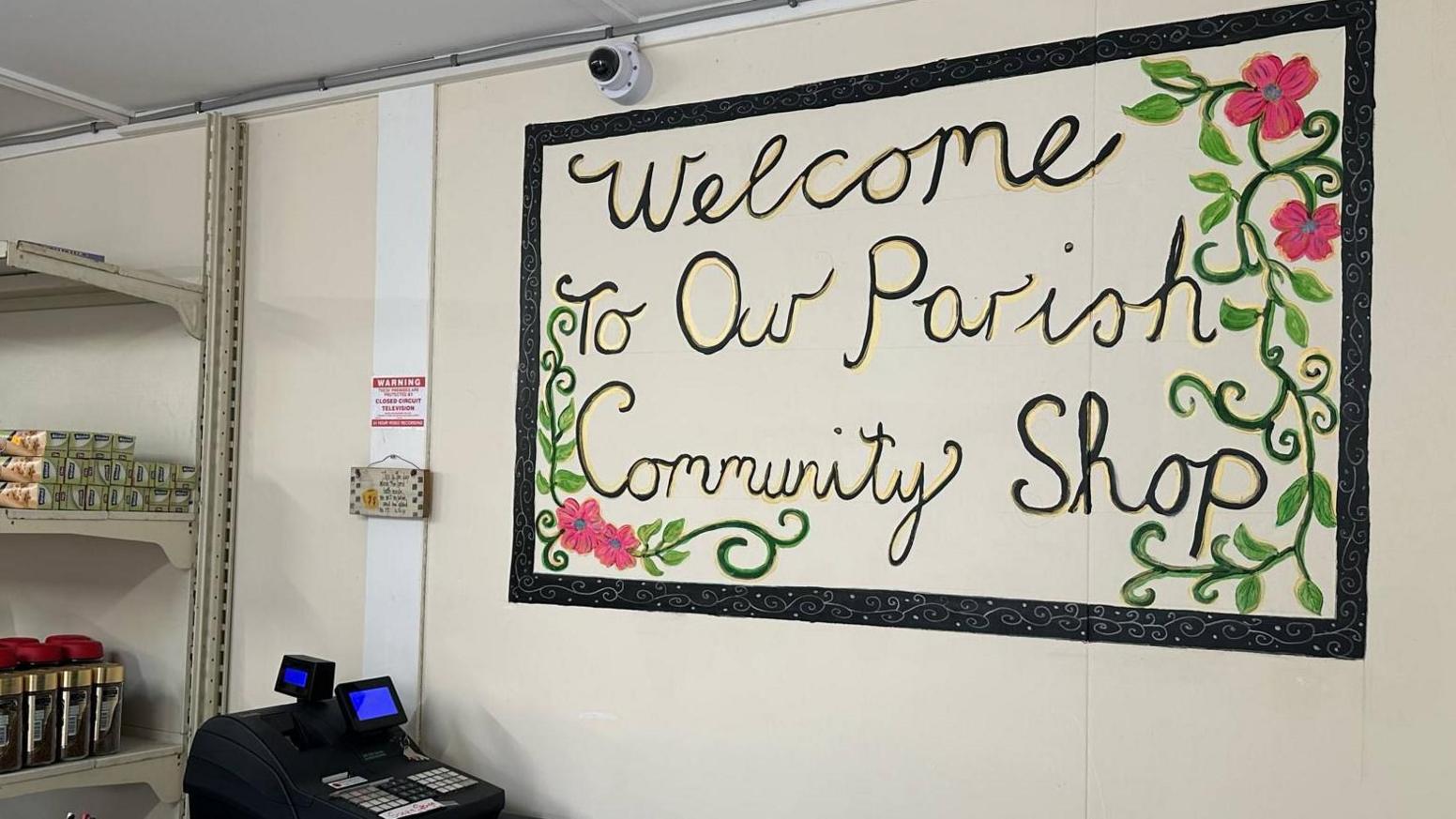 A 'Welcome to our parish community shop' sign which has a black border and green and pink flowers around it. There is a black till sitting on the bench and in the background you can see coffee pots and tinfoil sitting on shop shelves.