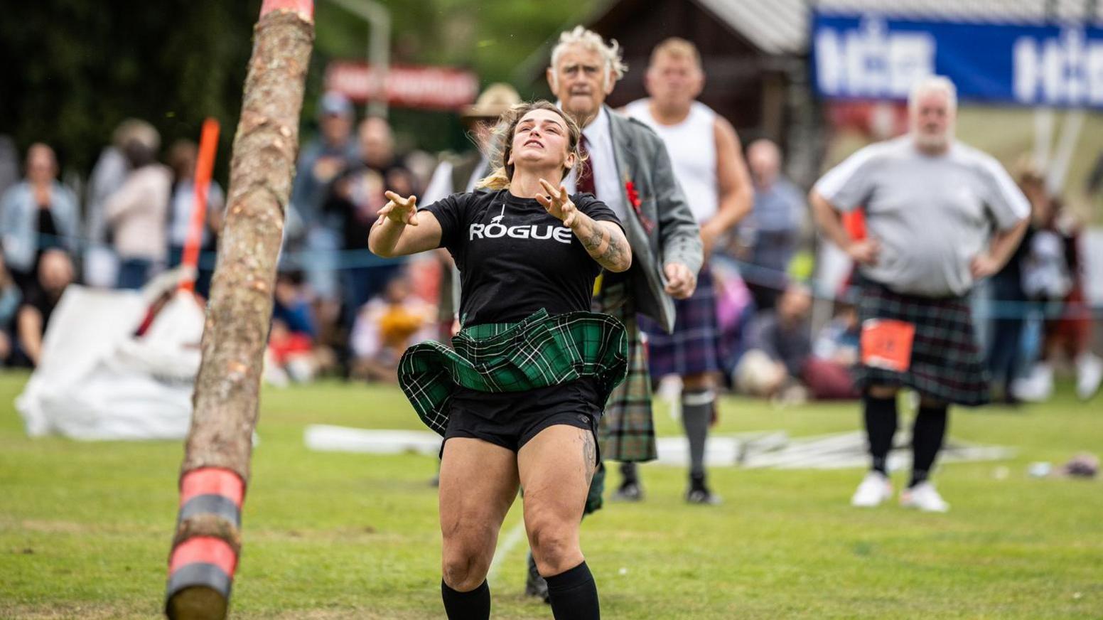 A female athlete leans back after tossing a caber. She is wearing a green tartan kilt and a black t-shirt with the word "rogue" in capitals. She is watched by a competition judge and male heavy athletes.