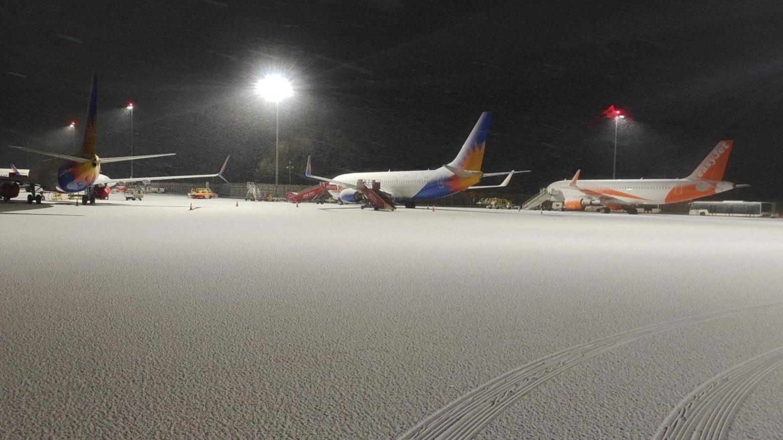 Three planes on snow-covered tarmac at Bristol Airport. A set of vehicle tracks can be seen in the foreground, but otherwise the snow is completely undisturbed.