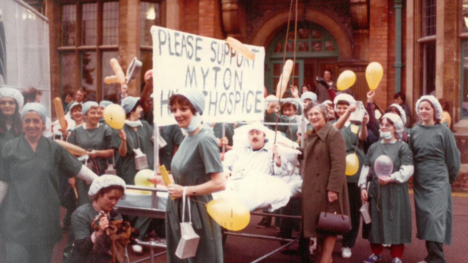 An old photo with a group of nurses around a hospital bed, with someone dressed as a patient in the bed. The nurses hold balloons and a big sign says 'Please Support Myton Hospice'